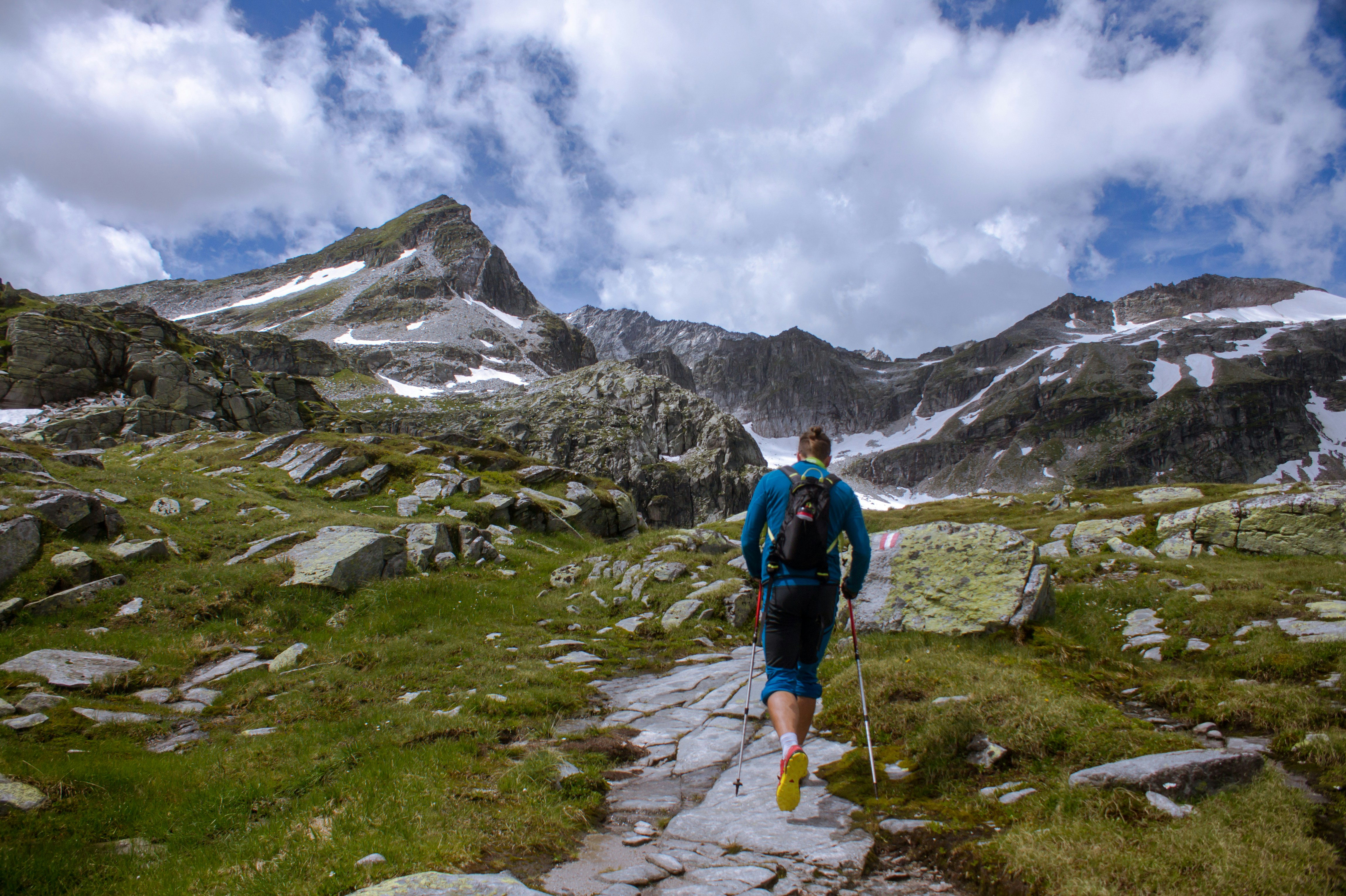 Hiker using trekking poles while walking along a rocky mountain trail surrounded by alpine scenery.
