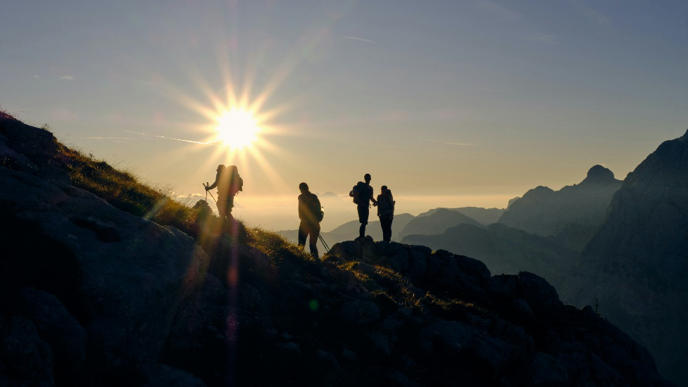 Group of hikers standing on a mountain ridge during sunrise with scenic alpine views in the background.