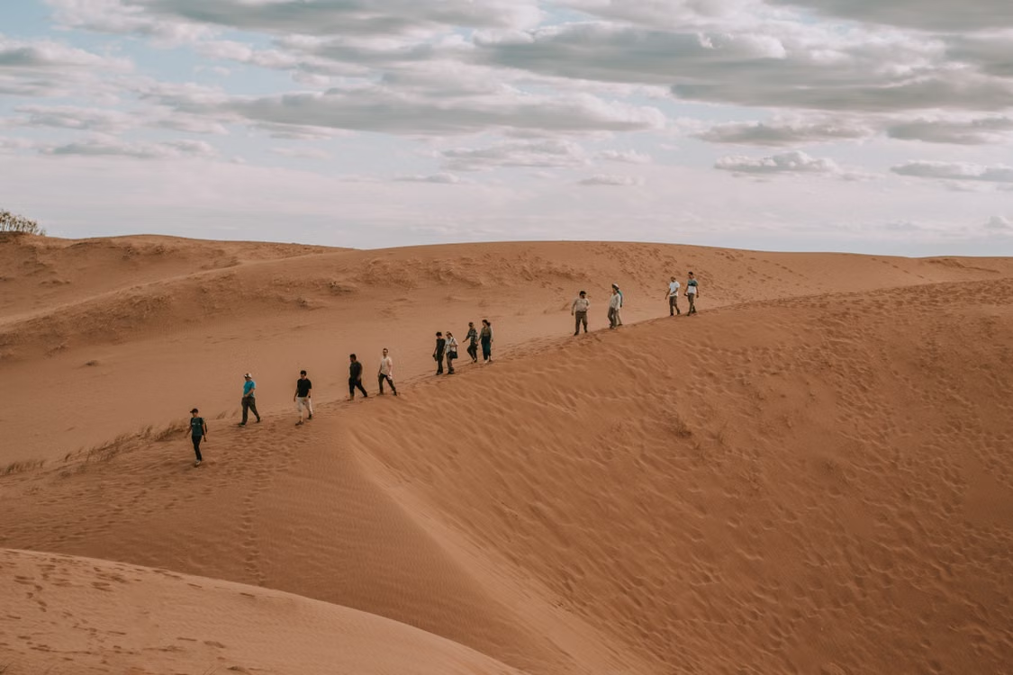 Line of hikers walking single file across large sand dunes under a cloudy sky