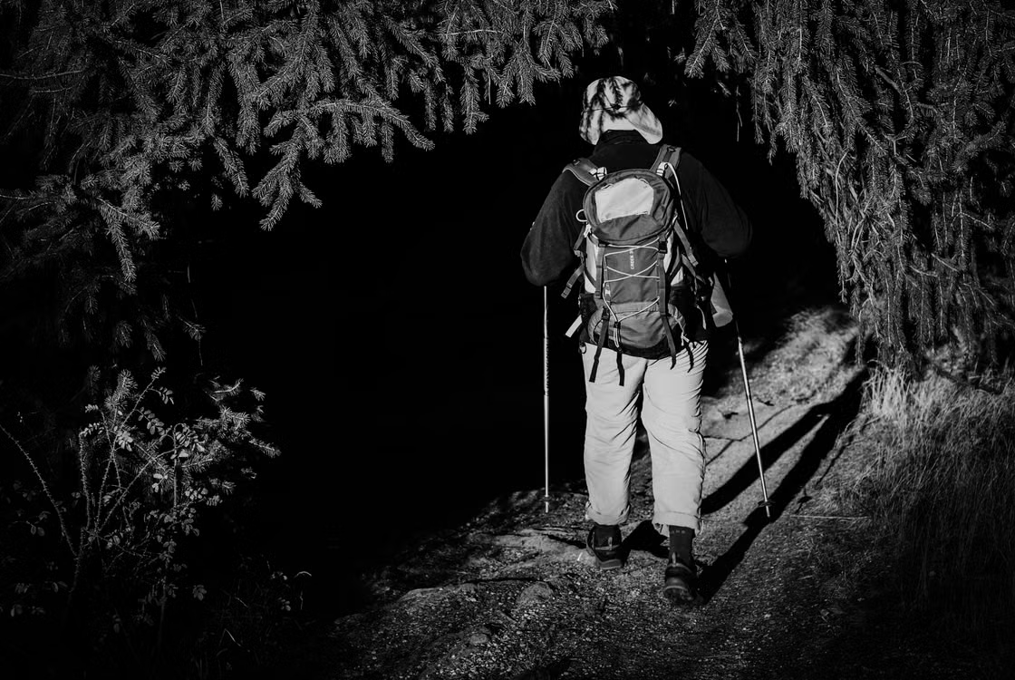 Hiker carrying a backpack and trekking poles on a dark forest trail at night.