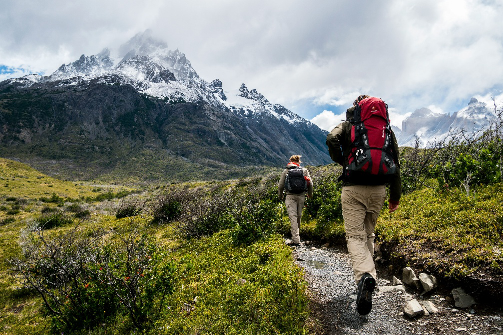 Two hikers carrying backpacks while walking along a mountain trail surrounded by rugged peaks and alpine terrain.